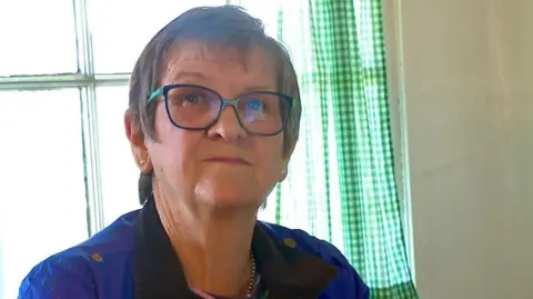 Eira Harris sitting in front of a window with green gingham curtains. She has short brown hair and is wearing glasses