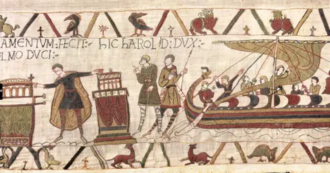 Bayeux Museum A wider crop of the same scene involving Harold, with him touching the boxes on the left of the image, and other figures nearby, with a ship next to them with people on, with medieval lettering and birds all around and fabric texture detail.