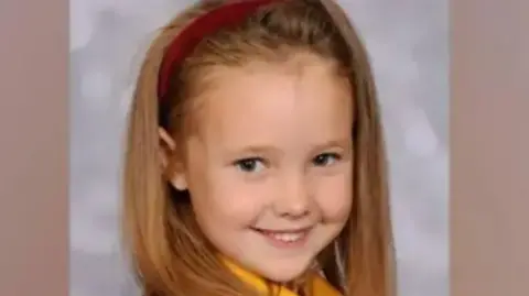 Family tells Elsie Dot Stancombe wearing a red headband and her school uniform, smiling while taking pictures.