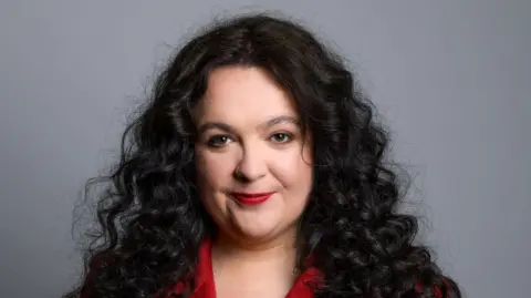 Ashley Storrie, a woman with long dark hair, wearing a red jacket. She is staring into the camera