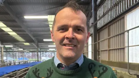 ALEX JAMES/BBC Jonny pictured looking into the camera and smiling. He's wearing a dark green knitted jumper with reindeer on it, and a light blue collared shirt and a navy tie. He's stood indoors inside the livestock market, a large warehouse or barn-type structure with corrugated walls and ceiling. 