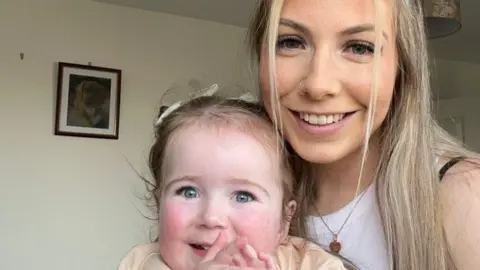 Sian Kennedy, who has long blonde hair, holding her little girl Sienna. Both are smiling at the camera.