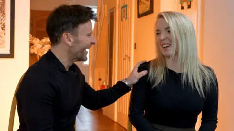 A man and woman gaze into each others eyes smiling, the man has his hand on the woman's shoulder