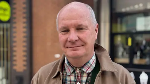 The image shows an older gentleman, the background is blurred and he's in the street. He's wearing a tartan shirt and a brown jacket. He's got short grey hair and he's smiling. 