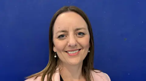 Ben Parker/BBC A head and shoulders image of Rachael Tecklenberg, wearing a pink blazer and standing in front of a deep blue background. She is smiling and looking into the camera. 
