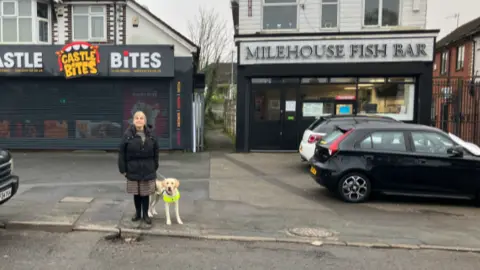 A woman is standing at the edge of a pavement in front of a pothole, with a dog in a luminous harness to her side. There are two takeaways behind her, and cars parked to the left and right
