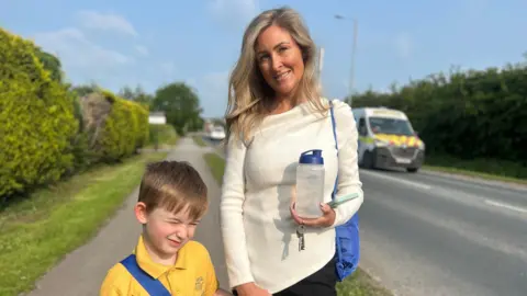 BBC News A woman is wearing a white top and is holding her son's water bottle and PE kit on the main road. Her son is holding her left hand, wearing a yellow polo shirt, which is his school uniform and wearing his book bag.
