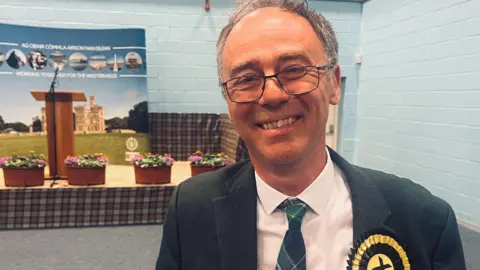 Alasdair Allan is pictured indoors. He has grey hair and is wearing glasses. He has a dark suit jacket over a white shirt and a tartan tie. He is wearing a yellow and black rosette.