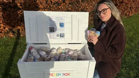 A woman wearing glasses and a brown coat kneels beside an open white wooden box labelled “Teddy Box.” Inside the box are many small, individually wrapped teddy bears.