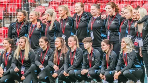20 women football players are pictured with eight in the front row crouching down and a further 10 standing behind them. They are wearing black tops and tracksuit bottoms and they are standing in a goalmouth with the netting visible behind them 