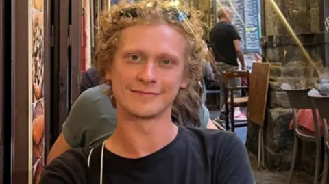 Syeva Bondarenko Syeva Bondarenko with curly blonde hair sits in a restaurant. He is wearing sunglasses on his head and a black top.