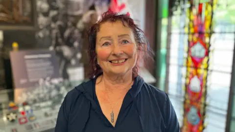 A woman, with short, curly red hair, smiling at the camera. She is wearing a navy top. 
