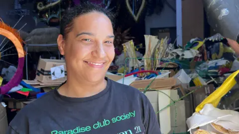 Lily-May Symonds/BBC A woman wearing a black t-shirt that says "paradise club society" in green font i smiling with carnival props scattered behind her.