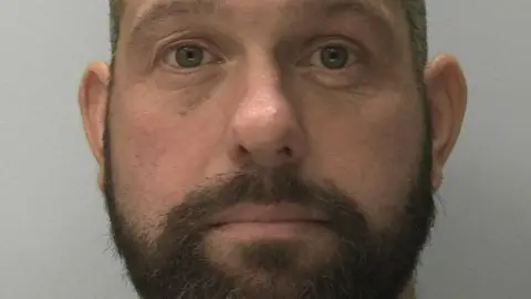 A police mugshot of James Desborough in front of a grey background. He is balding and has a brown beard