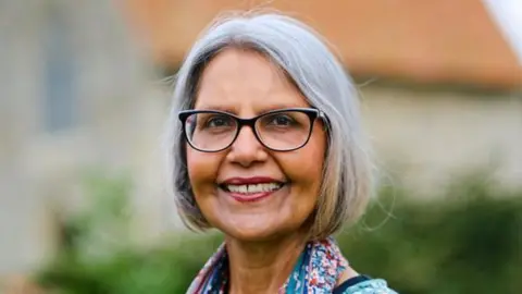 Kurshida Mirza: A grey-haired woman wearing dark-rimmed glasses and a blue, black and white patterned dress. 