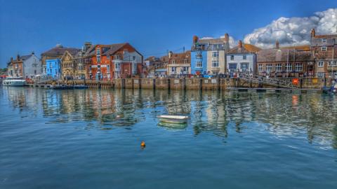 MONDAY - Weymouth Harbour on the south coast is a typical English Harbour, here colourful buildings can be seen on the quay and are reflected in the water. In the foreground there is a small white rowing boat. The sky is blue and the sun is shining