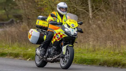 DERBYSHIRE BLOOD BIKES A motorcyclist is driving down the road. The motorcycle is covered in neon yellow ambulance stickers. The word 'blood' is on the front of the vehicle.