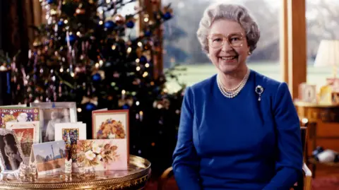 A woman wears glasses and pearls. She has short white hair and a brooch on a blue or purple dress. She is sitting and next to her is a gold circular table with cards on and a photograph. Behind them both iis a decorated Christmas tree. The room's windows can be seen and they view is of trees and grass. It is daytime and the woman has a wide-open smile.