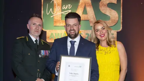 LAS Luke Rapela, stands holding a certificate and looks at the camera. To his left is a man with a blazer and medals on and to his right is a woman with long blonde hair and a yellow dress on. They are pictured at an awards ceremony