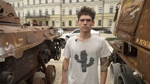 Oleksii between two military vehicles in Kyiv after his rescue. He has short brown hair and wears a white t-shirt with a picture of a cactus on it.