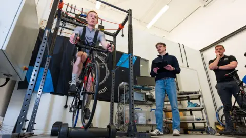 A cyclist wearing a grey top and black shorts, rides an exercise bike in a laboratory, as two researchers, one wearing a black polo neck shirt and blue jeans and holding a clip board, watch on.