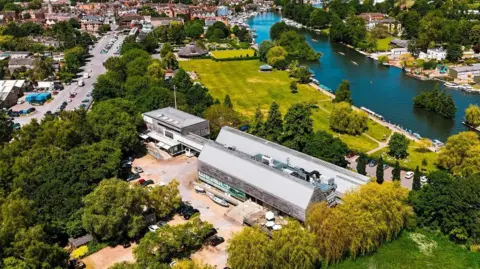 An aerial photo of the museum. It's a long thin building with a white roof, built near the river. There's trees and a large patch of grass next to it, with the town of Henley beyond.