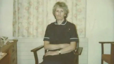 Nora sitting on a wooden chair, looking at the camera, in her navy and white nurses uniform.