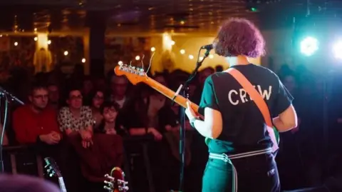 The back of a guitarist performing to a crowd of people. The performer is wearing a black T-shirt which says CREW in white writing.