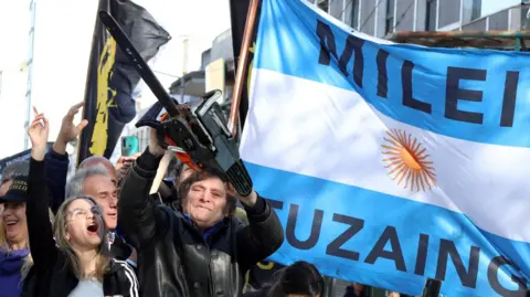 Reuters Javier Milei stands in a crowd during an election rally holding a chainsaw up to the air. An Argentinian flag with the word "Milei" is in the immediate background.
