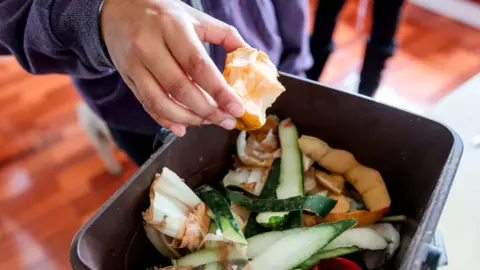 A person at home wearing a purple jumper drops an apple core into a small black food waste bin that is full of vegetable peelings.