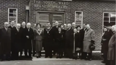 Friends of Woodston Library A group of people, in a black and white photo, standing outside a brick built library. They are all wearing coats and jackets. The image is slightly blurred. 
