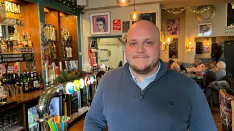 BBC A picture of Rob Moore in his pub. He is wearing a navy blue shirt and is smiling and looking at the camera.