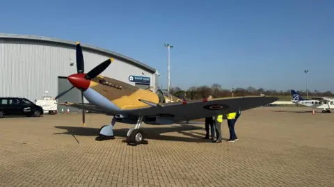 A brown Spitfire on the tarmac at outside aircraft hangers at the airport