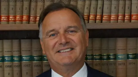 Paul Marshall has short brown hair and wears a black suit with a white shirt and red spotted tie.