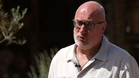 A bald man wearing a white shirt giving an interview outside