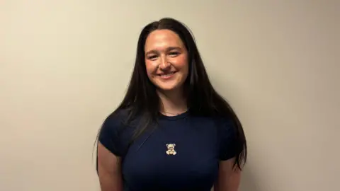 jessika is standing in front of a white wall. She is wearing a navy top with a teddy bear on it. She has long black hair. She also has a small chain around her neck.