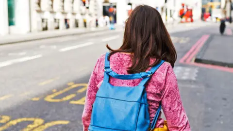 Getty Images A young woman with brown hair, a pink top and a blue packpack walks down a street