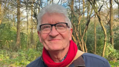 Verity Ure-Jones A man with black frame glasses. white hair, a red scarf and a blue coat smiles at the camera. Behind him is a woodland with green foliage and brown, spindly tree trunks.