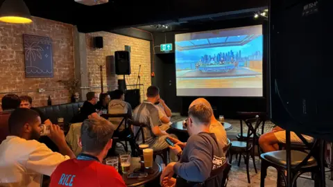 Copper Beech Brew Co showing the football on a large screen. People are sat around tables in the venue drinking beer and wearing football shirts. 
