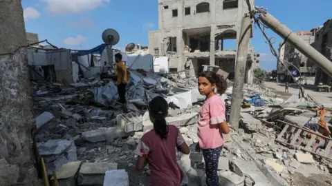 EPA Two Palestinian girls and two men inspect a destroyed home in the Magazi refugee camp, in the center of the Gaza Strip, following an Israeli air strike (14 August 2024)