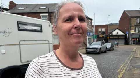 Simon Thake A woman in a white stripey top and grey hair tied back stands on a cobbled street. Some shops and bars can be seen in the background.