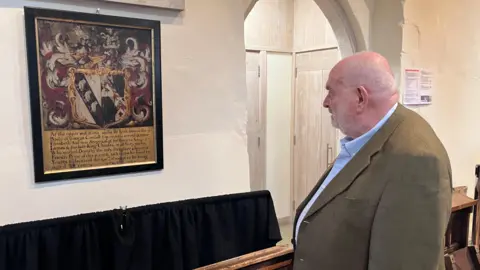 Sam Read/BBC A man looking at a framed plaque with a decorative coat of arms and text mounted on a stone wall, beside an arched doorway.