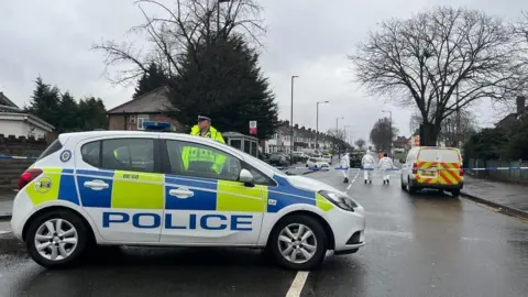 A police officer stood behind a police car on a single carriageway road with police tape across it and forensic officer dressed in white overalls walking up the road in the background