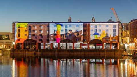 Adam Kenrick Huge projected images are reflected onto a brick building on the water. The images reflect back into the water in the evening's darkness. The projected pictures show a man in front of buildings and coloured statues.