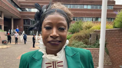 BBC Sandra Samuels, deputy lieutenant of the West Midlands. She is wearing a white blouse with a ruffled collar and a green suit and is standing next to the flagpole at the Civic Centre, with people behind her.