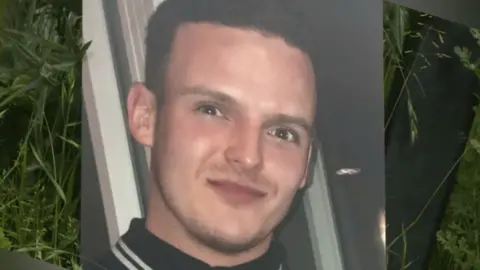 A large print out of a photo of Stephen Morrisson is propped up against foliage. He is wearing a black polo with white stripes on the collar. He has a slight smile and has dark brown hair.