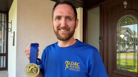 Aaron Beggs smiling at the camera holding a gold medal with a blue ribbon. He has brown hair and a beard and is wearing a blue tshirt with 'North Down Athletic Club' written on it in yellow.
