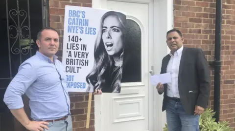 BBC Kristofer Deichler and Jatinder Kamra stand outside Catrin Nye's home, holding a poster with her face that includes the words "BBC's Catrin Nye's lies".