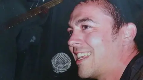 A side profile of a smiling Jon Casey dressed in a black shirt with a silver microphone near his mouth