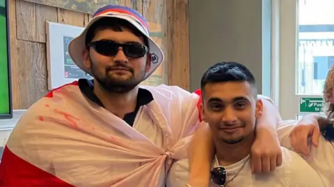 Jay Harrington Ashwin and his brother Jay smile at the camera. Ashwin has his arm around his brother's neck. Ashin wears sunglasses and a bucket hat with an flag wrapped around his body. Jay has short dark hair.